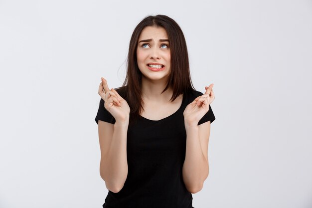 Belle jeune fille pleine d'espoir en t-shirt noir posant avec les doigts croisés sur le mur blanc