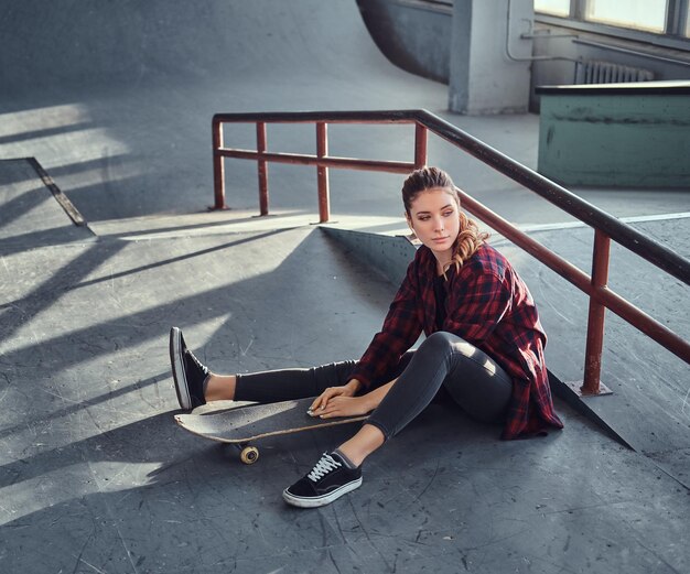 Une belle jeune fille portant une chemise à carreaux tenant une planche à roulettes tout en étant assise à côté d'un rail de broyage dans le skatepark à l'intérieur.