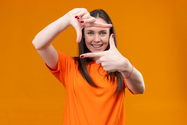 Belle jeune fille portant un t-shirt orange faisant cadre avec les mains et les doigts souriant joyeusement debout sur fond orange isolé