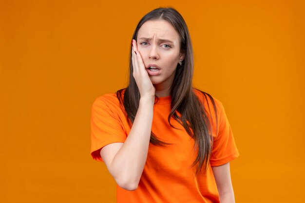 Belle jeune fille portant un t-shirt orange à la recherche de mal de toucher sa joue mal de dents debout sur fond orange isolé