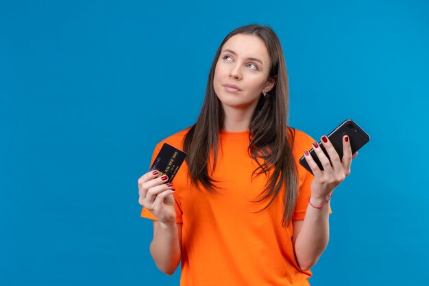 Belle jeune fille portant un t-shirt orange tenant un smartphone et une carte de crédit à la recherche avec une expression pensive pensant essayer de faire un choix debout sur fond bleu isolé