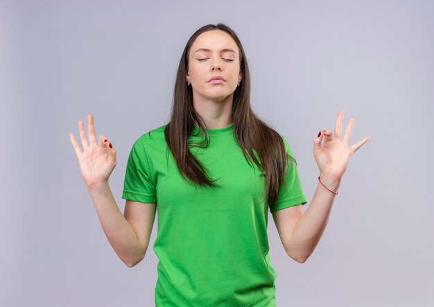Belle jeune fille portant un t-shirt vert se détendre avec les yeux fermant faisant le geste de méditation avec les doigts debout sur fond blanc isolé