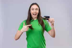 Photo gratuite belle jeune fille portant un t-shirt vert sorti et heureux tenant la carte de crédit pointant avec le doigt vers elle debout sur fond blanc isolé