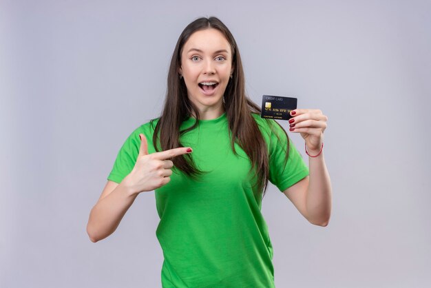 Belle jeune fille portant un t-shirt vert sorti et heureux tenant la carte de crédit pointant avec le doigt vers elle debout sur fond blanc isolé