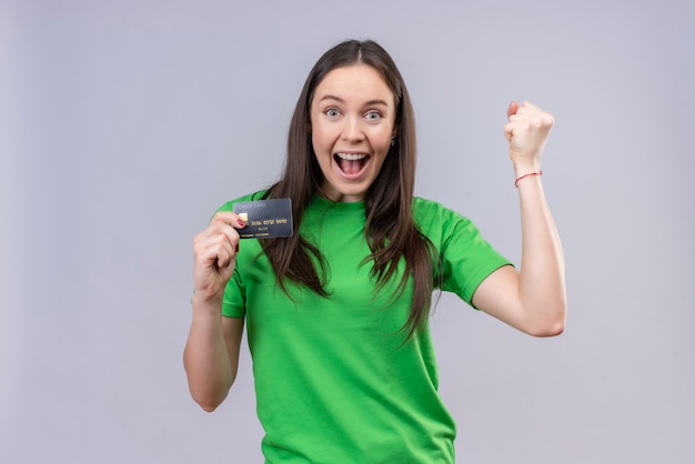 Belle jeune fille portant un t-shirt vert sorti et heureux tenant la carte de crédit souriant joyeusement levant le poing se réjouissant de son succès debout sur fond blanc isolé