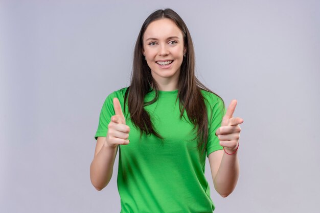 Belle jeune fille portant un t-shirt vert souriant joyeusement en pointant vers la caméra avec les deux mains debout sur fond blanc isolé