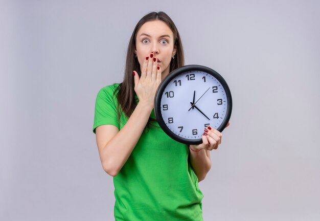 Belle jeune fille portant un t-shirt vert tenant une horloge à la bouche couvrant surpris et choqué avec la main debout sur fond blanc isolé