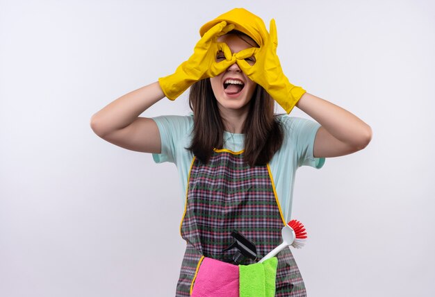 Belle jeune fille portant un tablier, une casquette et des gants en caoutchouc faisant des signes ok comme des jumelles avec les doigts regardant à travers les doigts qui sort la langue