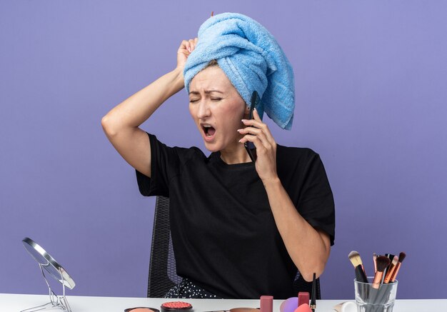 Une belle jeune fille regrettée est assise à table avec des outils de maquillage essuyant les cheveux dans une serviette parle au téléphone en mettant la main sur la tête isolée sur fond bleu