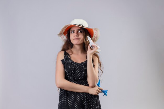 Belle jeune fille en robe à pois en chapeau d'été tenant des avions jouets à côté mécontent avec une expression sceptique sur le visage debout sur fond blanc