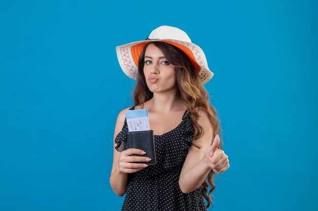 Belle jeune fille en robe à pois en chapeau d'été tenant des billets d'avion montrant les pouces vers le haut positif et heureux debout sur fond bleu