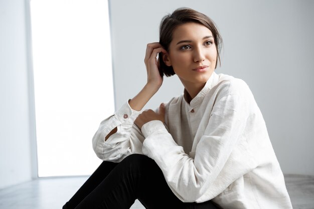 Belle jeune fille souriante assise sur le plancher sur le mur blanc.