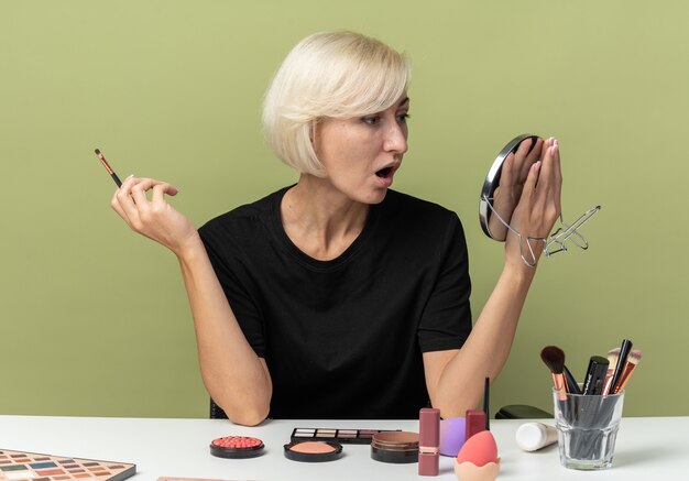 Belle jeune fille surprise est assise à table avec des outils de maquillage tenant un pinceau de maquillage avec miroir isolé sur fond vert olive