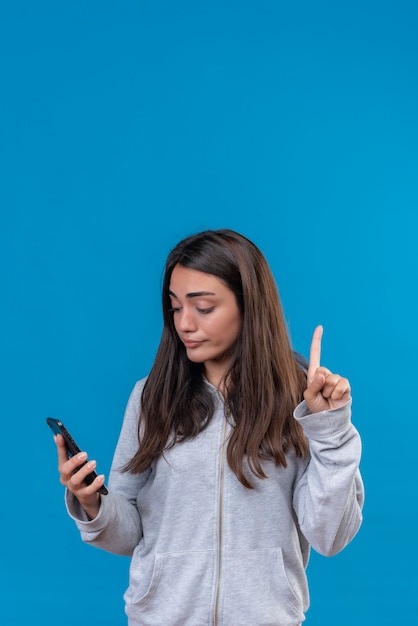 Belle jeune fille en sweat à capuche gris tenant le téléphone à la recherche de téléphone doigt pointé vers le haut debout sur fond bleu