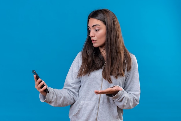 Belle jeune fille en sweat à capuche gris tenant le téléphone et regardant le téléphone avec une émotion surprise debout sur fond bleu