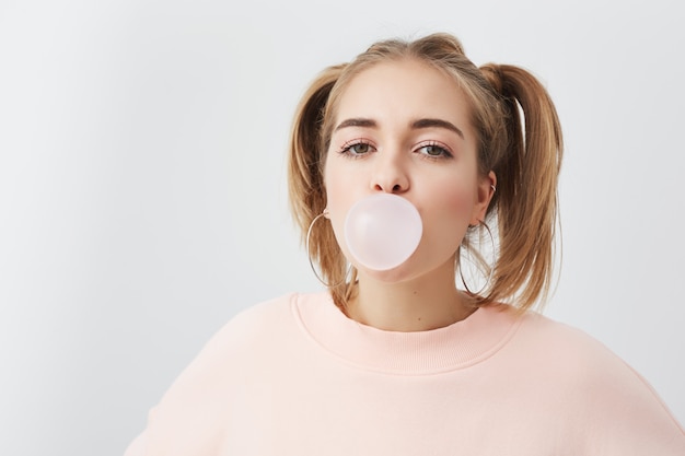 Photo gratuite belle, jolie et drôle jeune fille soufflant des bulles de chewing-gum. femme blonde avec deux queues de cheval sur la tête portant un sweat rose posant.