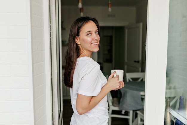 belle jolie femme aux cheveux noirs, sourire heureux portant un t-shirt blanc buvant le café du matin dans la cuisine. bonne journée ensoleillée.