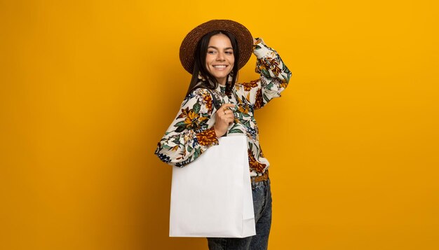 Belle jolie femme brune élégante posant isolé sur fond de studio jaune dans une tenue de vêtements de mode à la mode tenant un chemisier imprimé sac à provisions blanc et un chapeau souriant heureux accessoires