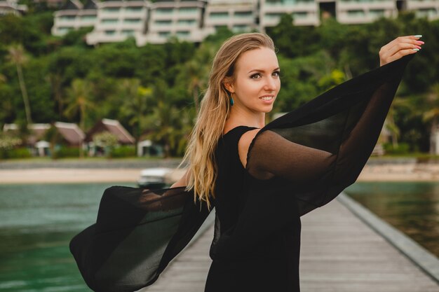 Belle jolie femme vêtue d'une robe noire posant sur la jetée dans un hôtel de luxe, vacances d'été, plage tropicale