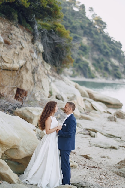 belle mariée aux cheveux longs en robe blanche avec son mari sur la plage près de grosses pierres