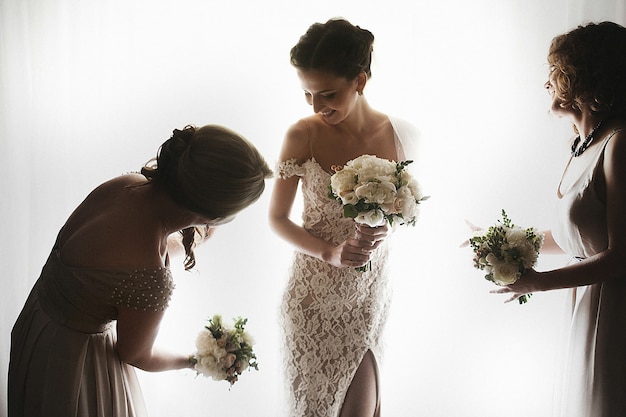 Photo gratuite belle mariée brune sexuelle en robe blanche avec demoiselles d'honneur près de la fenêtre