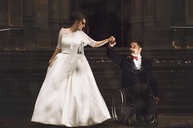 Belle mariée et le marié sur le fauteuil roulant dansent devant la vieille église catholique