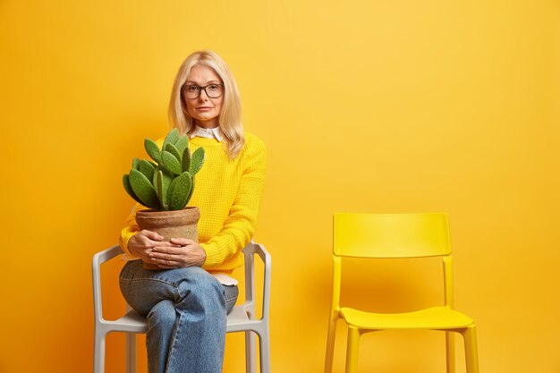 Belle mature femme européenne détient cactus en pot prend soin de la maison des fleurs vit seul pose sur une chaise confortable