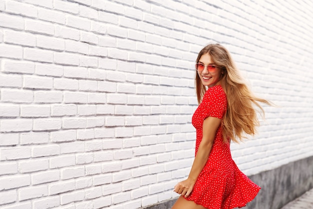 Belle petite amie blonde se retourne vers vous et souriant, explorant les rues de la ville en vacances d'été, l'air heureux, posant près de mur de briques blanches à l'extérieur