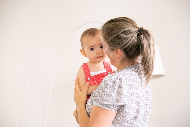 Belle petite fille assise sur les mains de maman. Mère blonde tenant sa fille, parler avec elle et souriant. Temps en famille, maternité et concept d'être à la maison