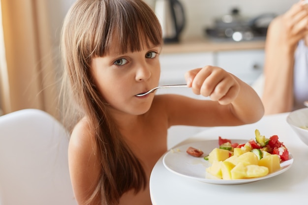 Belle petite fille aux cheveux noirs assise à table et mangeant des pommes de terre et une salade de légumes dans son assiette, regardant la caméra, posant à l'intérieur dans la cuisine.