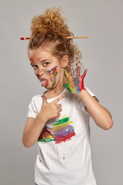 Belle petite fille ayant un pinceau dans sa jolie coupe de cheveux, vêtue d'un t-shirt blanc enduit. Elle pose avec des mains et des joues peintes, pointant sur sa main et montrant une langue, sur un dos gris