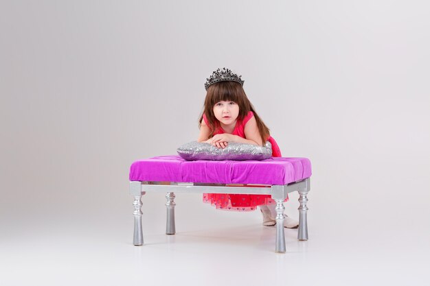 Belle Petite Fille Brune En Robe De Princesse Rose Avec Couronne Assise Sur Une Chaise Rose Et Jouant Avec Un Oreiller Bebe Mignon Photo Premium