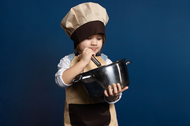 Photo gratuite belle petite fille chef cuisinier quelque chose dans une casserole. enfant de 5 ans concentré en tablier et chapeau fouettant les blancs d'œufs avec diligence tout en préparant la pâte à biscuit pour la pâtisserie. concept de cuisson