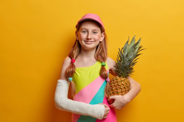 Belle petite fille en maillot de bain coloré et bonnet, pose avec de l'ananas contre le mur jaune, aime l'heure d'été et se repose bien, s'est cassé le bras après une chute de hauteur ou un sport dangereux