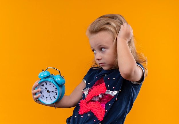 Photo gratuite une belle petite fille portant une chemise bleu marine tenant un réveil bleu tout en touchant sa tête sur un mur orange