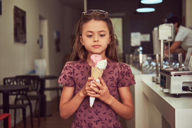 Une belle petite fille vêtue d'une robe à la mode tient une glace à la fraise, debout dans un salon de crème glacée.