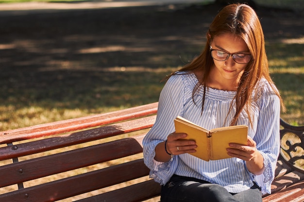 Belle photo d'une jeune fille dans une chemise bleue et avec des lunettes lisant un livre sur le banc