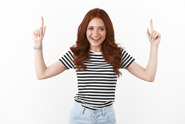 Photo gratuite belle rousse animée en t-shirt rayé riant joyeusement pointant les doigts levés vers le haut introduisez l'espace de copie supérieur promo regardez la caméra satisfaite recommandez une bonne promotion fond blanc