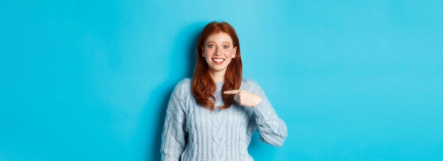 Photo gratuite belle rousse pointant vers elle-même et souriante heureuse d'être choisie debout en pull contre