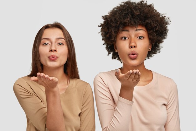 De belles jeunes femmes caucasiennes et afro-américaines soufflent un baiser d'air, expriment leur amour à d'autres personnes, disent au revoir à distance, vêtues d'une tenue décontractée, posent ensemble contre un mur blanc