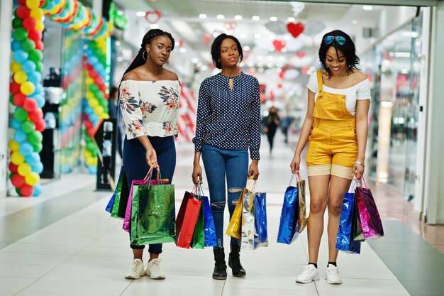 Belles trois filles afro-américaines bien habillées avec des sacs à provisions colorés dans un centre commercial