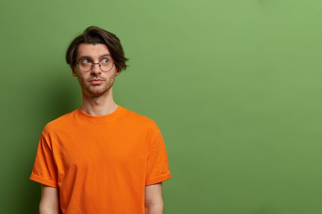 Besoin de penser. Un mec pensif avec une coupe de cheveux à la mode regarde sérieusement de côté, réfléchit au plan ou à la décision, porte des lunettes rondes et un t-shirt orange, isolé sur un espace vide de mur vert