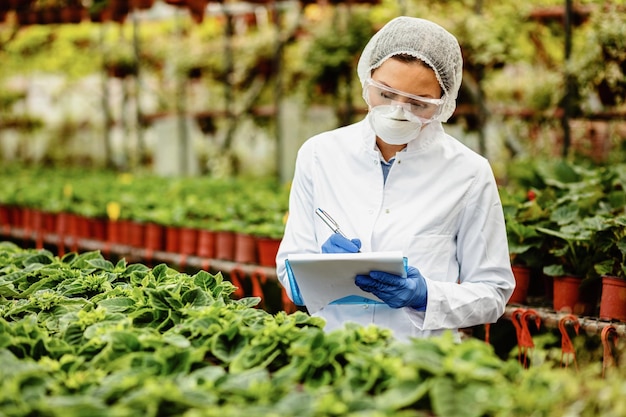 Biotechnologiste femelle inspectant des plantes en pot dans une pépinière et écrivant des notes dans le presse-papiers