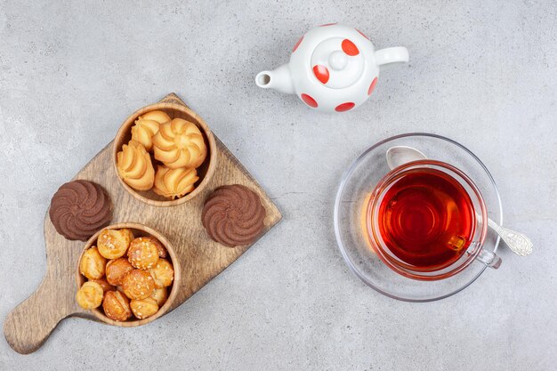 Biscuits dans des bols et sur planche de bois, avec une tasse de thé et une petite théière sur une surface en marbre