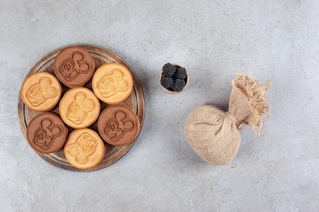 Biscuits décorés sur planche de bois à côté d'un sac et un petit bol de mûres sur fond de marbre. Photo de haute qualité