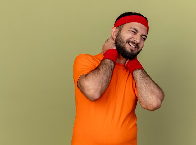 Blessé jeune homme sportif portant bandeau et bracelet a attrapé le cou douloureux isolé sur fond vert olive