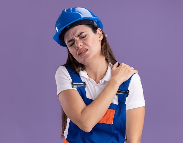Blessée aux yeux fermés, une jeune femme de construction en uniforme a attrapé une épaule douloureuse isolée sur un mur violet