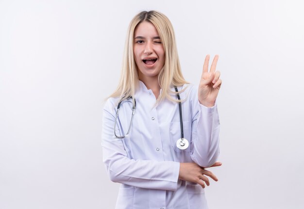 Blinked joyeuse médecin jeune fille blonde portant un stéthoscope et une robe médicale en orthèse dentaire montrant le geste de paix sur un mur blanc isolé