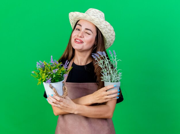 Blinked smiling belle fille de jardinier en uniforme portant chapeau de jardinage tenant et traversant des fleurs en pot de fleurs isolé sur vert