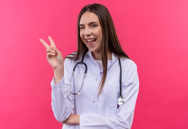 Blinked young doctor girl wearing stethoscope medical gown montrant la langue et le geste de paix sur mur rose isolé